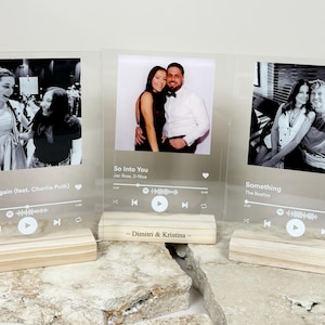 May include: Three clear acrylic photo frames with a wooden base. Each frame features a black and white photo and a Spotify-style music player graphic with the song title and artist name. The frames are personalized with the names "Dimitri & Kristina" and the song titles "See You Again (feat. Charlie Puth)", "So Into You", and "Something".