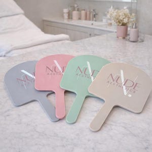Op de afbeelding: Vier handwaaiers in pastelkleuren: grijs, roze, mintgroen en beige. Elke waaier heeft een handvat en de woorden "NUDE AESTHETICS" zijn in een complementaire kleur gedrukt. De waaiers liggen op een marmeren oppervlak.