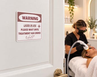 May include: A rectangular white sign with maroon text and borders mounted on a door. The sign reads WARNING LASER IN USE PLEASE WAIT UNTIL TREATMENT HAS ENDED and features a small star icon. The text Enchanted Skin & Beauty is below.