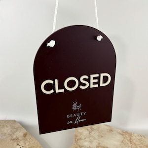 May include: A dark burgundy arch-shaped sign hanging by a white cord. The sign features the white 3D text "CLOSED" and the white printed logo and text "BEAUTY in bloom" with a small floral line art icon centered below the main word.