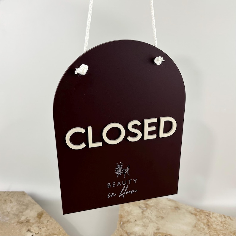 May include: A dark burgundy arch-shaped sign hanging by a white cord. The sign features the white 3D text "CLOSED" and the white printed logo and text "BEAUTY in bloom" with a small floral line art icon centered below the main word.