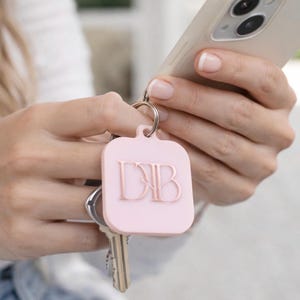 May include: A light pink keychain with the initials "DB" in a serif font. The keychain is square with rounded corners and is attached to a silver keyring with keys. The keychain is held in a person's hands, along with a smartphone.