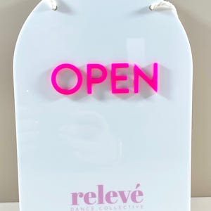 May include: An arched white acrylic sign featuring the word OPEN in raised neon pink lettering. Below the text, the words relev&eacute; DANCE COLLECTIVE are printed in a smaller pink font. The sign includes two white cords at the top for hanging.