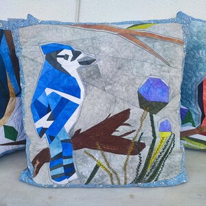 May include: A bluebird pillow with a blue and white patterned design, sitting on a gray background with brown branches and green leaves. The pillow is made of fabric and has a blue border.