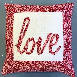 May include: Red and white quilted pillow with a white square in the center featuring the word "love" in red fabric. The pillow has a floral pattern.