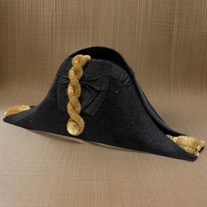 Op de afbeelding: Een zwarte bicorne met gouden galonversiering. De hoed heeft een decoratief ontwerp aan de voorkant en een gouden knoop aan de zijkant.
