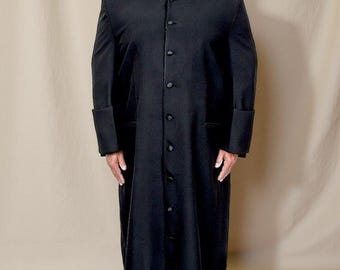 May include: A black clergy robe with a white collar and button-down front. The long-sleeved robe has folded cuffs and is worn by a person. The background is a neutral beige color.