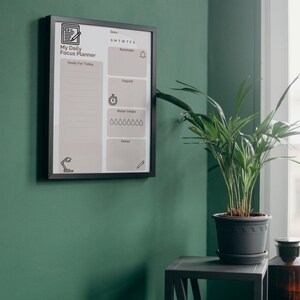 Op de afbeelding: Een ingelijste "My Daily Focus Planner" hangt aan een groene muur. De planner heeft secties voor doelen, herinneringen en waterinname. Een potplant staat op een kleine tafel naast de planner.