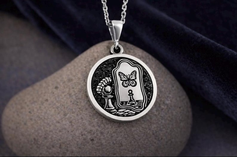 May include: A circular silver pendant on a silver chain featuring an engraved design of a caterpillar on a chess piece looking into a mirror that reflects a butterfly. The background of the engraving is dark and textured.