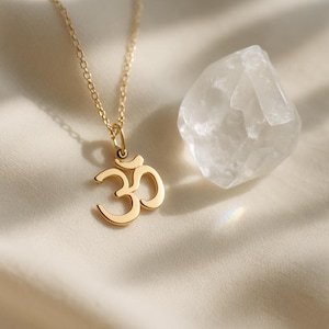 Peut inclure: Un collier doré avec un pendentif symbole Om. Le pendentif a la forme du symbole Om. Un cristal de quartz clair est à côté du collier sur une surface crème. La chaîne du collier est délicate.