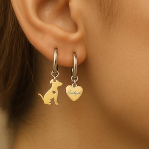 Può includere: Un paio di orecchini a cerchio color oro. Un orecchino presenta una silhouette di cane con un cuore ritagliato, e l'altro ha un ciondolo a forma di cuore con la scritta "Luckycb" in corsivo.