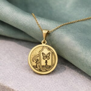 May include: A gold-toned circular pendant necklace featuring an engraved design of a chess piece, a butterfly, and a mirror. The pendant hangs from a delicate gold-toned chain, showcasing intricate line art details on its polished surface.
