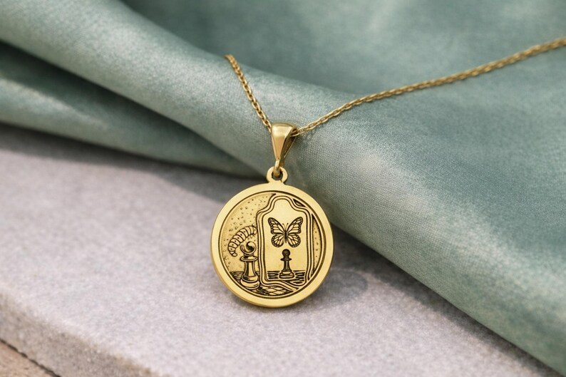 May include: A gold-toned circular pendant necklace featuring an engraved design of a chess piece, a butterfly, and a mirror. The pendant hangs from a delicate gold-toned chain, showcasing intricate line art details on its polished surface.