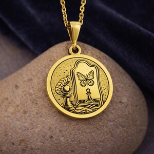 May include: A gold-toned circular pendant necklace featuring an engraved illustration of a caterpillar perched on a chess pawn, looking into a mirror that reflects a butterfly and a chess pawn. The pendant hangs from a matching gold chain.