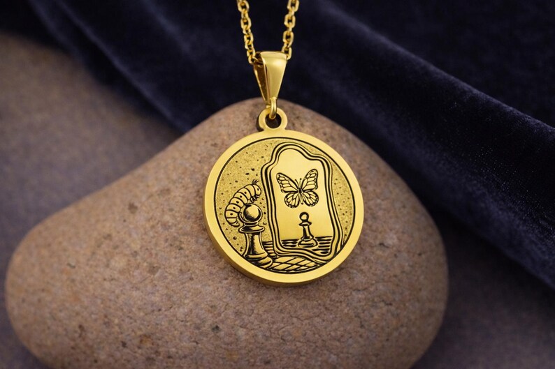 May include: A gold-toned circular pendant necklace featuring an engraved illustration of a caterpillar perched on a chess pawn, looking into a mirror that reflects a butterfly and a chess pawn. The pendant hangs from a matching gold chain.