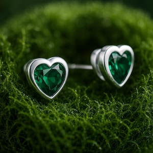 Può includere: Orecchini a lobo a forma di cuore con un'intensa pietra verde incastonata in metallo color argento. Gli orecchini sono esposti su un letto di muschio verde, evidenziandone il colore e il design.