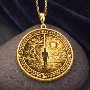 May include: A gold-toned circular pendant on a matching chain. The pendant features an embossed design of a person standing between a stormy sea with lightning and a calm sea with a sun. The text AMOR FATI is engraved at the top.