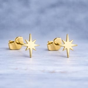 Könnte beinhalten: Ein Paar goldfarbene Stern-Ohrstecker. Jeder Ohrring hat ein Stern-Design mit acht Spitzen. Die Ohrringe werden auf einer hellen Oberfläche mit verschwommenem Hintergrund präsentiert.