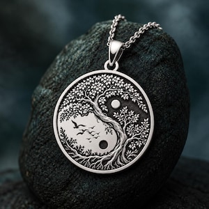 May include: A circular silver pendant on a chain featuring an embossed yin and yang design. The imagery depicts a tree with detailed leaves and roots, with a flock of birds flying in the negative space and two small circular accents.