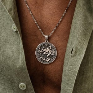 Op de afbeelding: Een zilverkleurige ronde hanger aan een ketting met een reliëfafbeelding van een figuur die over een wateroppervlak leunt om de eigen reflectie te bekijken, omringd door een decoratieve rand, hangend aan een dunne zilverkleurige ketting.