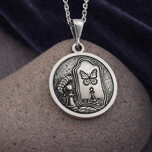 May include: A circular silver pendant on a chain featuring an embossed design of a caterpillar crawling toward a chess pawn. The pawn is reflected in a mirror as a butterfly. The metal has a dark, oxidized finish in the recessed areas.