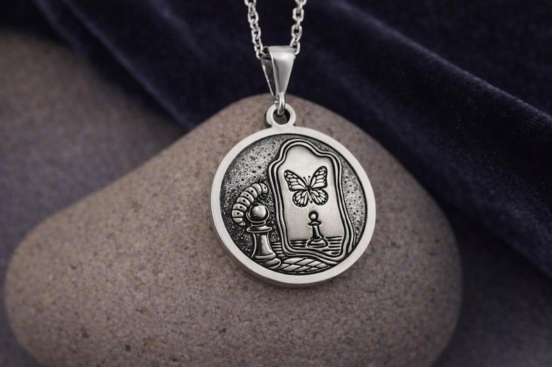 May include: A circular silver pendant on a chain featuring an embossed design of a caterpillar crawling toward a chess pawn. The pawn is reflected in a mirror as a butterfly. The metal has a dark, oxidized finish in the recessed areas.