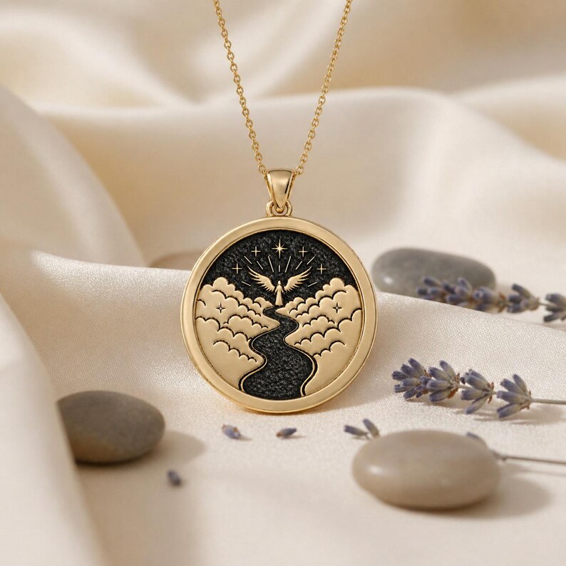 May include: A gold-toned circular pendant necklace featuring a black textured inlay. The design depicts a central winged figure standing above a winding path surrounded by stylised clouds and four-pointed stars under a dark night sky.