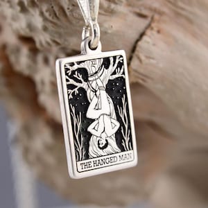 Könnte beinhalten: Ein rechteckiger silberner Anhänger mit einer schwarz-weißen Gravur der Tarotkarte Der Gehängte. Das Motiv zeigt eine Figur, die kopfüber an einem Baum hängt, mit der römischen Ziffer XII und dem Text THE HANGED MAN.