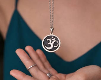 May include: Silver-toned pendant necklace featuring a circular pendant with the Om symbol in black. The necklace is suspended from a delicate chain. The pendant is held in an open hand, with rings on the fingers. The background is a teal-colored top.