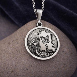 May include: A circular silver pendant on a chain featuring an engraved design of a caterpillar next to a chess pawn, with a mirror reflecting a butterfly and a chess piece. The metal has a polished finish with dark recessed details.