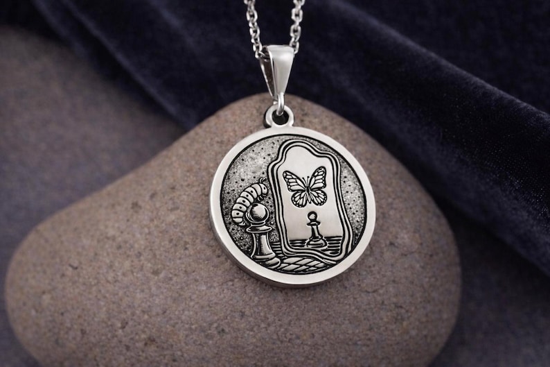 May include: A circular silver pendant on a chain featuring an engraved design of a caterpillar next to a chess pawn, with a mirror reflecting a butterfly and a chess piece. The metal has a polished finish with dark recessed details.