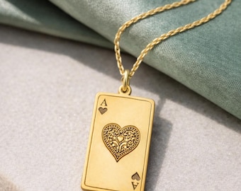 May include: Gold-colored pendant necklace featuring an Ace of Hearts playing card design. The pendant has a detailed heart in the center and the card's suit symbols. The necklace is displayed on a light gray surface with a green fabric background.