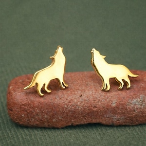 May include: A pair of gold-colored wolf-shaped stud earrings. The earrings are in the shape of a howling wolf, with the head tilted upwards. The earrings are displayed on a reddish-brown stone against a green background.