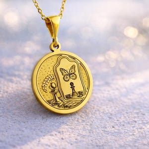 May include: A gold-toned circular pendant necklace featuring an engraved design of a caterpillar, a chess pawn, and a butterfly reflected in a mirror. The pendant hangs from a delicate gold chain, showcasing intricate line work and a polished finish.