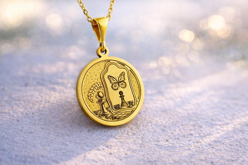 May include: A gold-toned circular pendant necklace featuring an engraved design of a caterpillar, a chess pawn, and a butterfly reflected in a mirror. The pendant hangs from a delicate gold chain, showcasing intricate line work and a polished finish.
