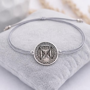 May include: A silver-colored bracelet featuring a round pendant with an hourglass design. The pendant is surrounded by a Celtic knot pattern. The bracelet is strung on a gray cord and rests on a gray pillow.