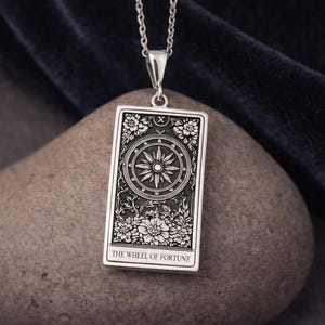May include: A silver pendant necklace with a rectangular tarot card design. The card shows a wheel with floral and star details, and the words "THE WHEEL OF FORTUNE" are at the bottom. The pendant hangs from a delicate silver chain.