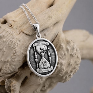 May include: A silver oval pendant necklace featuring an hourglass design. The top bulb contains a skull, and the bottom bulb contains a tree. The text MEMENTO MORI is engraved at the top and MEMENTO VIVERE is engraved at the bottom of the pendant.