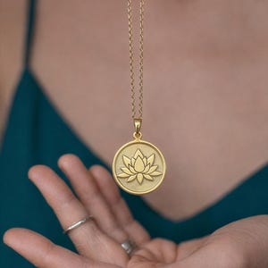 May include: Gold-colored pendant necklace featuring a circular charm with a lotus flower design. The pendant hangs from a delicate chain. The necklace is displayed against a teal background.