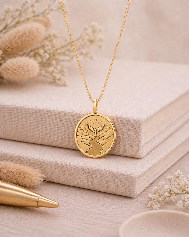 May include: A gold-toned necklace featuring a circular pendant with an embossed design of a winged figure standing at the end of a winding path amidst clouds and stars. The pendant hangs from a delicate gold-toned chain with a polished finish.