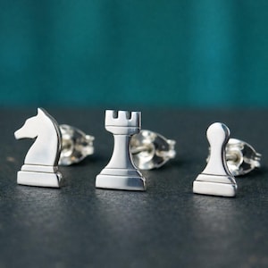 May include: Silver chess piece stud earrings. The set includes a knight, rook, and pawn. The earrings are displayed on a dark surface with a teal background. The pieces are highly detailed.