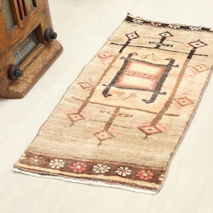 May include: A rectangular woven rug featuring a beige field with a central geometric motif in dark brown and soft pink. The border displays a dark brown band with repeating floral patterns in red and white, complemented by diamond-shaped accents.