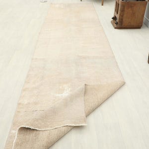 K&ouml;nnte beinhalten: Ein beigefarbener rechteckiger Teppich mit einem Used-Look-Muster ist teilweise auf einem hellen Holzboden ausgerollt. Die R&auml;nder des Teppichs sind ausgefranst, und die Gesamt&auml;sthetik ist neutral und strukturiert. Eine dekorative Vase und ein Holzschrank befinden sich im Hintergrund.