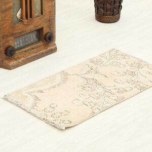 May include: A rectangular area rug featuring a light beige base color with a faded, intricate floral pattern in muted gray tones. The rug has short, frayed fringe edges on the short sides and a low-pile texture throughout the surface.