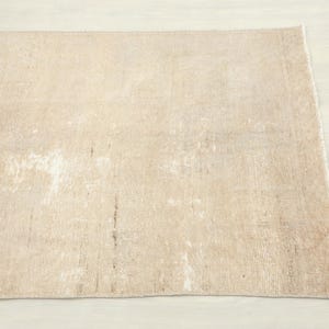 K&ouml;nnte beinhalten: Ein rechteckiger, beigefarbener Teppich mit einem Used-Look. Der Teppich hat ein verblasstes, abgenutztes Aussehen mit helleren Flecken und dunkleren Streifen, was auf Alter oder einen Vintage-Stil hindeutet. Die Kanten sind leicht ausgefranst, was seinen rustikalen Charme unterstreicht.