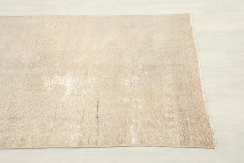 K&ouml;nnte beinhalten: Ein rechteckiger, beigefarbener Teppich mit einem Used-Look. Der Teppich hat ein verblasstes, abgenutztes Aussehen mit helleren Flecken und dunkleren Streifen, was auf Alter oder einen Vintage-Stil hindeutet. Die Kanten sind leicht ausgefranst, was seinen rustikalen Charme unterstreicht.