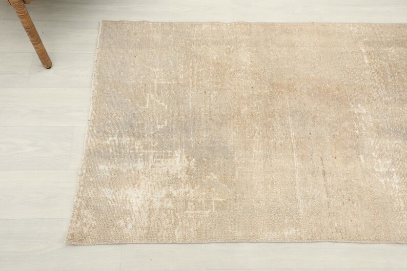 K&ouml;nnte beinhalten: Ein rechteckiger, beigefarbener Teppich mit einem abgenutzten, Vintage-Aussehen. Der Teppich hat ein verblasstes, gesprenkeltes Muster mit helleren und dunkleren Bereichen, wodurch ein abgenutzter Look entsteht. Der Teppich liegt auf einem hellen Holzboden, ein Teil eines Korbstuhls ist sichtbar.