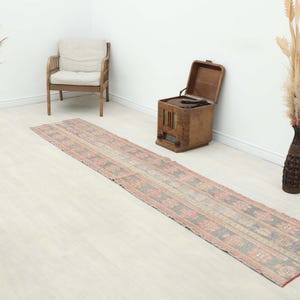 Può includere: Un lungo tappeto rettangolare dall'aspetto sbiadito e vintage. Il tappeto presenta un motivo ripetuto di forme geometriche in tonalità tenui di grigio, rosso e beige. Il tappeto è posizionato su un pavimento in legno chiaro, con una sedia in legno e oggetti decorativi sullo sfondo.