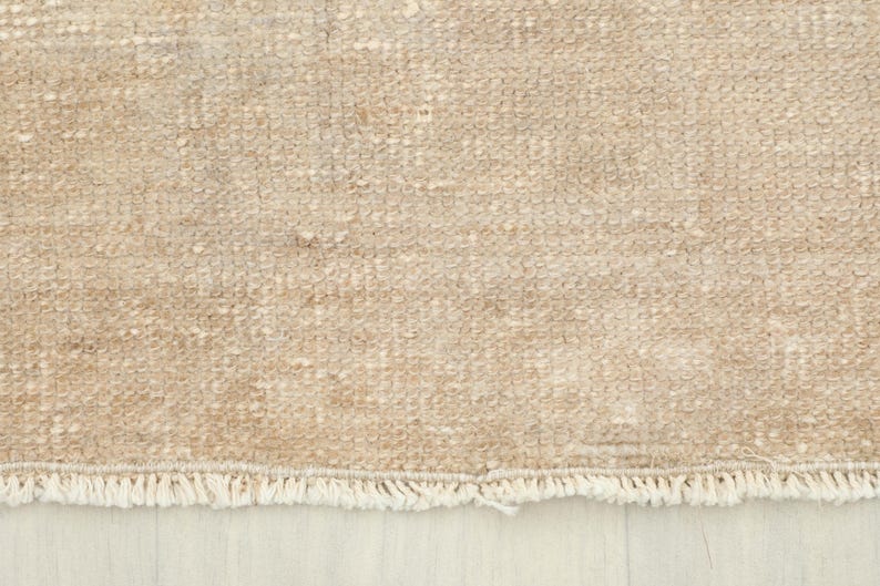 K&ouml;nnte beinhalten: Nahaufnahme eines beigefarbenen Teppichs mit strukturierter Oberfl&auml;che. Der Teppich hat ein gewebtes Muster und einen Fransenbesatz am unteren Rand. Die Farbe ist ein neutrales Beige, passend f&uuml;r verschiedene Einrichtungsstile.