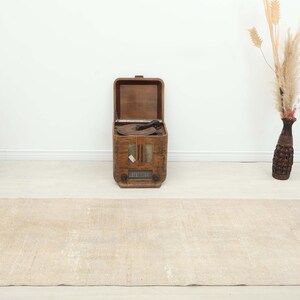 K&ouml;nnte beinhalten: Ein beigefarbener rechteckiger Teppich mit einem Used-Look. Ein offenes, vintage Holzradio steht aufrecht in der Mitte, mit einem Korbstuhl links. Eine dekorative Vase mit getrockneten Pflanzen befindet sich rechts.